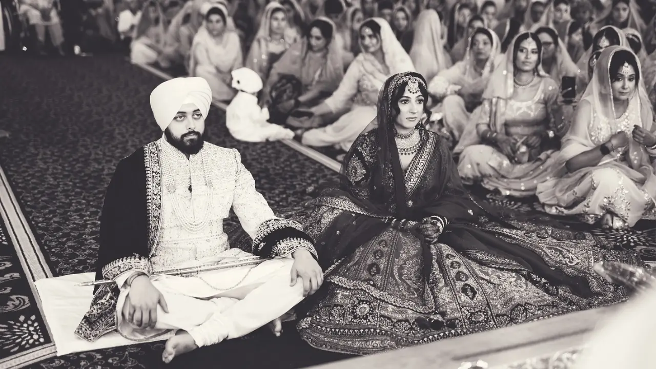 A Sikh couple sitting during their wedding ceremony, surrounded by women dressed in traditional attire, beautifully captured in black and white by Royal Bindi Photography.