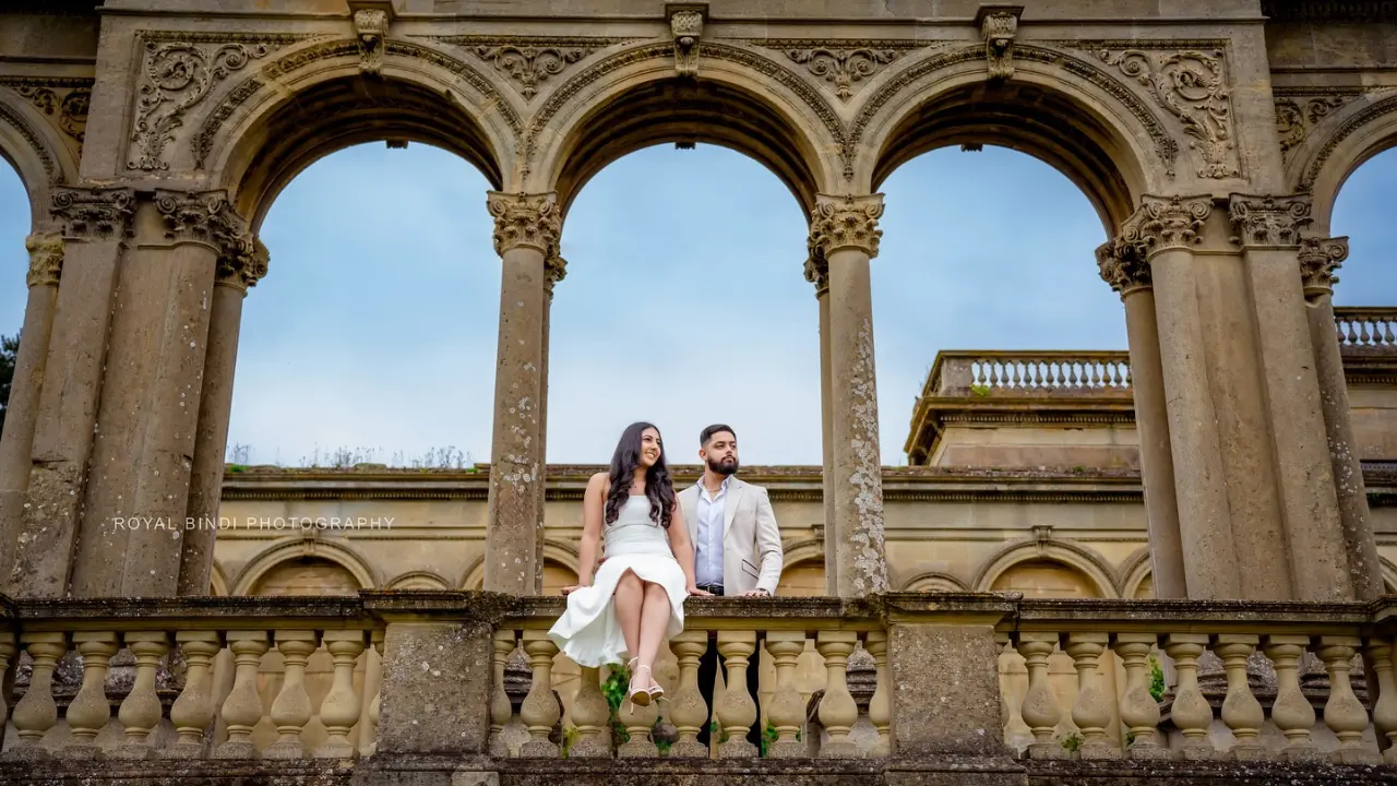 A couple poses elegantly on a stone balcony with ornate arches and historic architecture in the background, captured by Royal Bindi Photography.